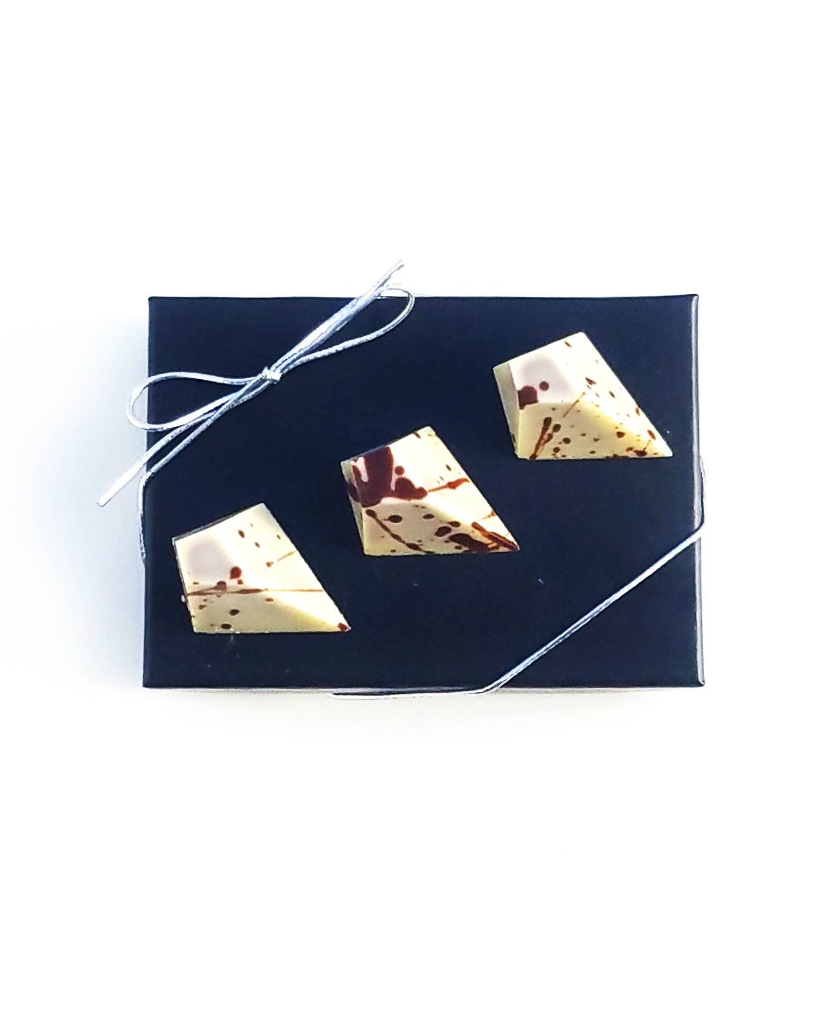 Three geometric Cafe au Lati truffles from Pyroclastic Chocolate sit atop a black box, tied with a silver bow, on a white background.